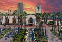 Union Station Train Station Surrounded By Office Buildings, Parked Cars, Tall Lush Green Palm Trees And Plants And People Walking On The Sidewalk With Powerful Clouds At Sunset In Los Angeles 