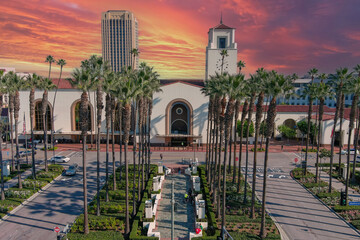 Wall Mural - Union Station train station surrounded by office buildings, parked cars, tall lush green palm trees and plants and people walking on the sidewalk with powerful clouds at sunset in Los Angeles 