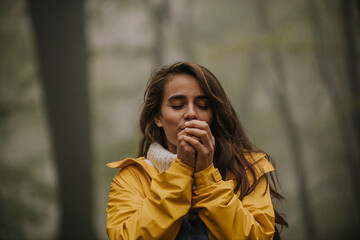 Smiling grateful woman in a yellow coat standing in the forest and enjoying the fresh mountain air. A woman is Blowing into her Hands on a Cold Winter day