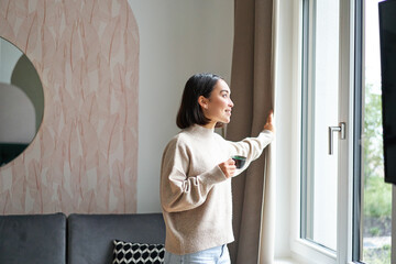 Wall Mural - Morning. Happy asian woman drinking espresso at home, looking outside window and smiling, standing in cozy living room feeling warm