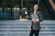© Strelciuc - Portrait of a happy businesswoman carrying laptop computer while walking to office. Empty space. Beautiful brunette outdoor. Mobile phone communication. Happy
