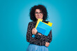 © Strelciuc - Front view portrait of cheerful young brunette business woman wearing eyeglasses isolated on blue background studio. Hold notebooks, looking at camera.
