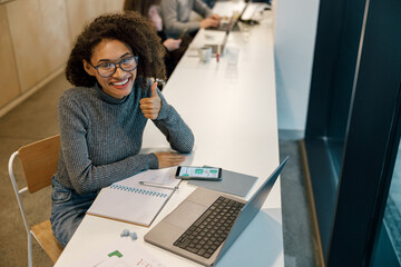 Wall Mural - Smiling woman freelancer working laptop and showing thumb up while sitting in cozy coworking