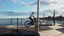 Senior Man Riding A Mountain Bike Along A Beach Walkway On A Sunny Day With Some Clouds. Lateral Camera Tracking