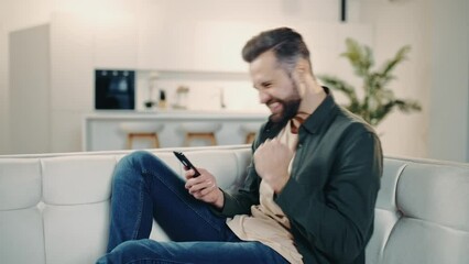 Joyful lucky attractive caucasian man in casual wear, sitting on a couch in cozy living room, using his smart phone, reading good message or news, gesturing with fist, smiling. Digital technology