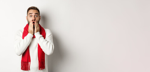 Christmas holidays and celebration concept. Handsome bearded guy in red scarf and sweater looking shocked, impressed with advertisement, white background