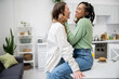 © LIGHTFIELD STUDIOS - happy and lesbian african american woman sitting on dining table and hugging cheerful girlfriend.