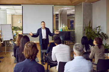 Wall Mural - Business trainer in suit and shirt is greeting group of adult business people students in modern office spreading his arms asides. People sitting at desks backs to camera. Adult education concept.