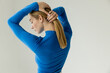 back view of blonde woman in blue long sleeve shirt making ponytail hairstyle isolated on grey.
