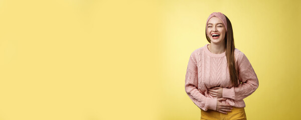 Amused and entertained charming young european female student laughing till belly hurts from giggle smiling holding palms on stomach, having fun enjoying watching funny comedy over yellow wall