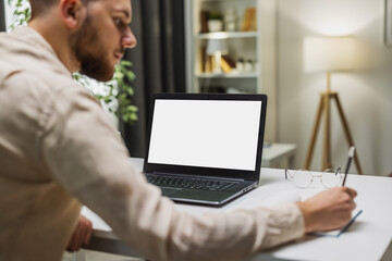 Man sitting at desk in front of blank screen laptop and writing in notebook, distant education concept