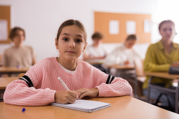 Wall Mural - Young girl sitting at desk in classroom and looking at camera.