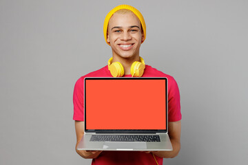 Wall Mural - Young IT man of African American ethnicity 20s he wear pink t-shirt yellow hat headphones hold use work on laptop pc computer with blank screen workspace area isolated on plain grey background studio.