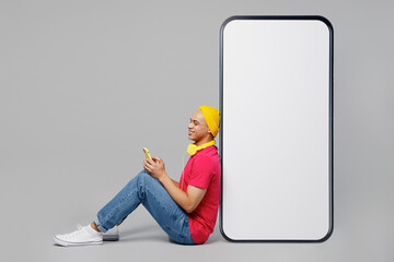 Wall Mural - Full body young man of African American ethnicity wear pink t-shirt yellow hat headphones sit big huge blank screen mobile cell phone with area use smartphone isolated on plain grey background studio.