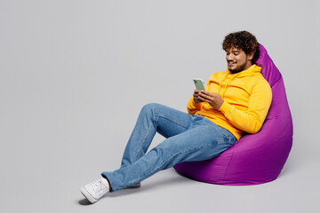 Wall Mural - Full body smiling young Indian man 20s he wearing casual yellow hoody sit in bag chair hold in hand use mobile cell phone isolated on plain grey background studio portrait. People lifestyle portrait.