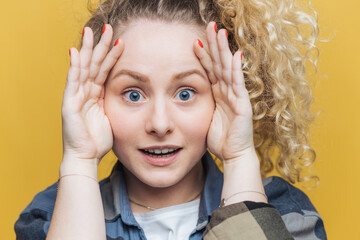 Wall Mural - Close up shot of emotional cute female with healthy skin and blue eyes, keeps hands on head, looks scrupulously and surprisingly at camera, notices something, isolated over yellow background