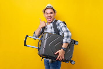 Wall Mural - Excited traveler tourist Asian man in casual clothes hat backpack holding suitcase bag, shows thumb up gesture on yellow background. Passenger travel abroad weekends getaway Air flight journey concept