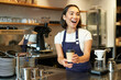 © Mix and Match Studio - Portrait of smiling asian girl barista, giving out order in cafe, inviting guest to pick up takeaway order near counter, holding takeaway cup of coffee