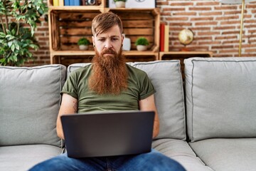 Wall Mural - Redhead man with long beard using laptop sitting on the sofa at the living room thinking attitude and sober expression looking self confident