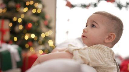 Poster - Adorable toddler sitting on sofa by christmas tree with serious expression at home