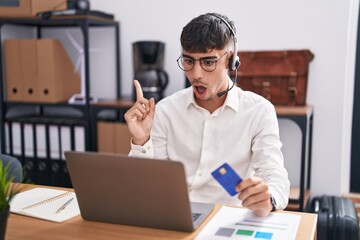 Wall Mural - Young hispanic man working using computer laptop holding credit card surprised pointing with finger to the side, open mouth amazed expression.