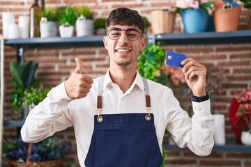 Canvas Print - Young hispanic man working at florist shop holding credit card smiling happy and positive, thumb up doing excellent and approval sign