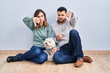 Poster - Young hispanic couple sitting on the floor with dog looking unhappy and angry showing rejection and negative with thumbs down gesture. bad expression.