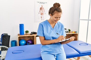 Canvas Print - Young beautiful hispanic woman physiotherapist smiling confident writing on document at rehab clinic