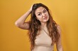 Young hispanic woman standing over yellow background confuse and wonder about question. uncertain with doubt, thinking with hand on head. pensive concept.