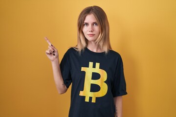 Poster - Blonde caucasian woman wearing bitcoin t shirt pointing with hand finger to the side showing advertisement, serious and calm face