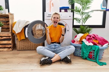 Wall Mural - Young blonde woman doing laundry sitting by washing machine celebrating crazy and amazed for success with arms raised and open eyes screaming excited. winner concept