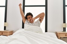 Young Hispanic Man Waking Up Stretching Arms Sitting On The Bed At Bedroom.
