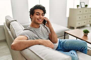 Wall Mural - Young hispanic man talking on the smartphone sitting on the sofa at home.