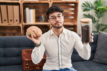 Wall Mural - Hispanic man with beard working at therapy office holding brain and coffee beans smiling looking to the side and staring away thinking.