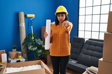 Sticker - Young hispanic woman standing on new home with blueprints pointing with finger to the camera and to you, confident gesture looking serious