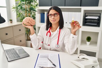 Wall Mural - Young hispanic doctor woman holding brain and cbd oil smiling looking to the side and staring away thinking.