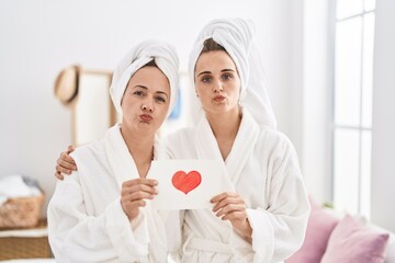 Wall Mural - Middle age woman and daughter wearing bath robe holding heart card looking at the camera blowing a kiss being lovely and sexy. love expression.