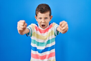 Wall Mural - Young caucasian kid standing over blue background angry and mad raising fists frustrated and furious while shouting with anger. rage and aggressive concept.