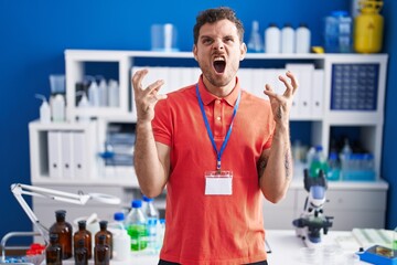 Sticker - Young hispanic man working at scientist laboratory crazy and mad shouting and yelling with aggressive expression and arms raised. frustration concept.