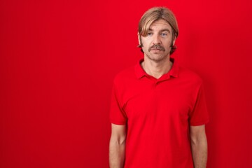 Poster - Caucasian man with mustache standing over red background relaxed with serious expression on face. simple and natural looking at the camera.