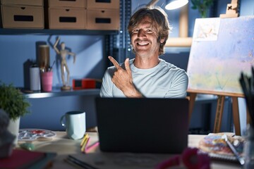 Poster - Middle age man sitting at art studio with laptop at night cheerful with a smile of face pointing with hand and finger up to the side with happy and natural expression on face