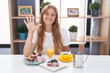 Sticker - Young caucasian woman eating pastries t for breakfast showing and pointing up with fingers number five while smiling confident and happy.