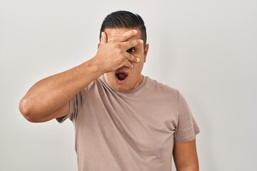 Wall Mural - Hispanic young man standing over white background peeking in shock covering face and eyes with hand, looking through fingers with embarrassed expression.