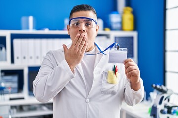 Canvas Print - Hispanic young man working at scientist laboratory holding id card covering mouth with hand, shocked and afraid for mistake. surprised expression