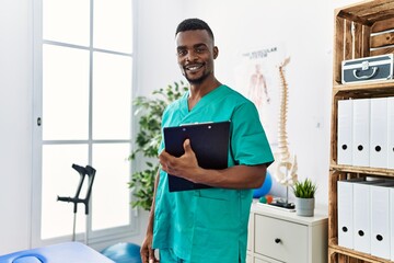 Wall Mural - Young african physiotherapist man working at pain recovery clinic looking positive and happy standing and smiling with a confident smile showing teeth