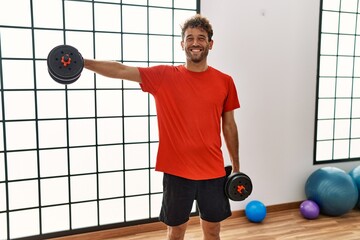 Wall Mural - Young hispanic man smiling confident training using dumbbells at sport center