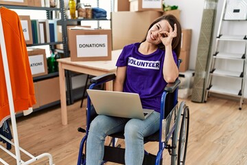 Wall Mural - Young brunette woman as volunteer on donations stand sitting on wheelchair smiling happy doing ok sign with hand on eye looking through fingers