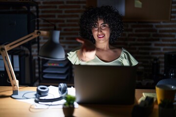 Wall Mural - Young brunette woman with curly hair working at the office at night smiling cheerful offering palm hand giving assistance and acceptance.