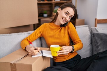 Wall Mural - Young caucasian woman talking on smartphone writing on notebook at new home