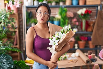 Wall Mural - Asian young woman at florist shop holding bouquet of flowers scared and amazed with open mouth for surprise, disbelief face